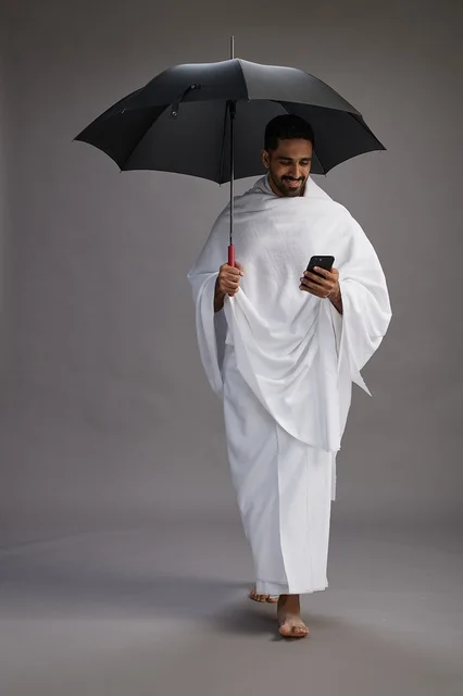 A portrait of an Arab Saudi Gulf pilgrim in Ihram clothing holding an umbrella, going to perform the rituals of Hajj and Umrah, using a mobile phone, full body, gray background.