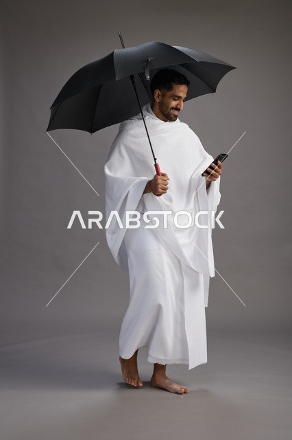 بورتريه لحاج  عربي سعودي  خليجي بملابس الإحرام يحمل مظله, الذهاب لاداء مناسك الحج والعمره, استخدام الهاتف المحمول  كامل الجسم, خلفيه رماديه