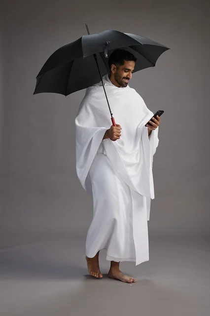 A portrait of an Arab Saudi Gulf pilgrim in Ihram clothing holding an umbrella, going to perform the rituals of Hajj and Umrah, using a mobile phone, full body, gray background.