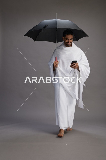 بورتريه لحاج  عربي سعودي  خليجي بملابس الإحرام يحمل مظله, الذهاب لاداء مناسك الحج والعمره, استخدام الهاتف المحمول  كامل الجسم, خلفيه رماديه