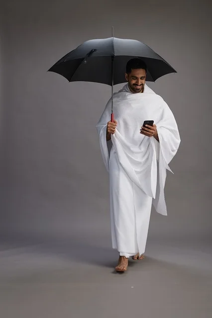 A portrait of an Arab Saudi Gulf pilgrim in Ihram clothing holding an umbrella, going to perform the rituals of Hajj and Umrah, using a mobile phone, full body, gray background.