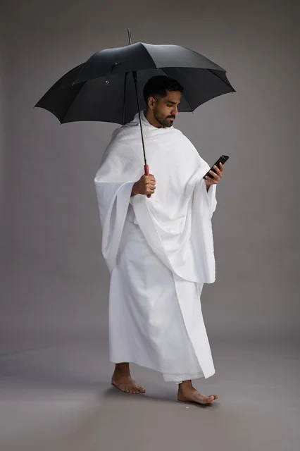A portrait of an Arab Saudi Gulf pilgrim in Ihram clothing holding an umbrella, going to perform the rituals of Hajj and Umrah, using a mobile phone, full body, gray background.