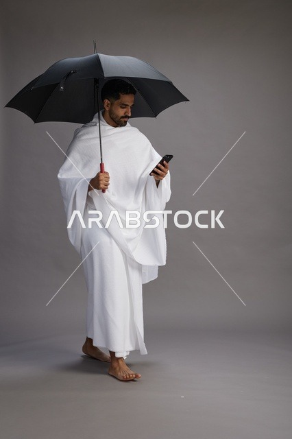بورتريه لحاج  عربي سعودي  خليجي بملابس الإحرام يحمل مظله, الذهاب لاداء مناسك الحج والعمره, استخدام الهاتف المحمول  كامل الجسم, خلفيه رماديه