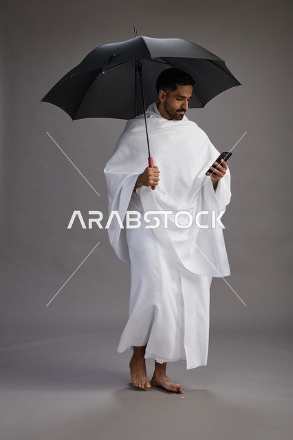 بورتريه لحاج  عربي سعودي  خليجي بملابس الإحرام يحمل مظله, الذهاب لاداء مناسك الحج والعمره, استخدام الهاتف المحمول  كامل الجسم, خلفيه رماديه
