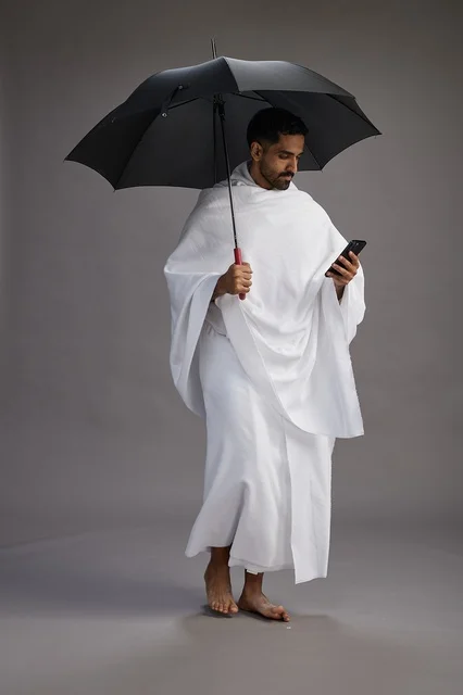 A portrait of an Arab Saudi Gulf pilgrim in Ihram clothing holding an umbrella, going to perform the rituals of Hajj and Umrah, using a mobile phone, full body, gray background.