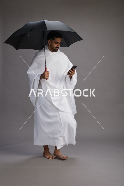 بورتريه لحاج  عربي سعودي  خليجي بملابس الإحرام يحمل مظله, الذهاب لاداء مناسك الحج والعمره, استخدام الهاتف المحمول  كامل الجسم, خلفيه رماديه