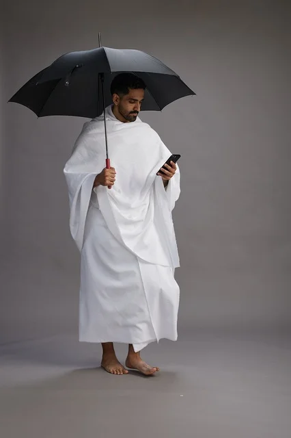 A portrait of an Arab Saudi Gulf pilgrim in Ihram clothing holding an umbrella, going to perform the rituals of Hajj and Umrah, using a mobile phone, full body, gray background.