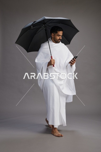بورتريه لحاج  عربي سعودي  خليجي بملابس الإحرام يحمل مظله, الذهاب لاداء مناسك الحج والعمره, استخدام الهاتف المحمول  كامل الجسم, خلفيه رماديه