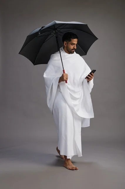 A portrait of an Arab Saudi Gulf pilgrim in Ihram clothing holding an umbrella, going to perform the rituals of Hajj and Umrah, using a mobile phone, full body, gray background. A portrait of an Arab Saudi Gulf pilgrim in Ihram clothing holding an umbrella, going to perform the rituals of Hajj and Umrah, using a mobile phone, full body, gray background.
