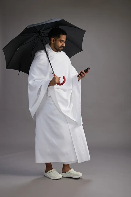 A portrait of an Arab Saudi Gulf pilgrim in Ihram clothing holding an umbrella, going to perform the rituals of Hajj and Umrah, using a mobile phone, full body, gray background.