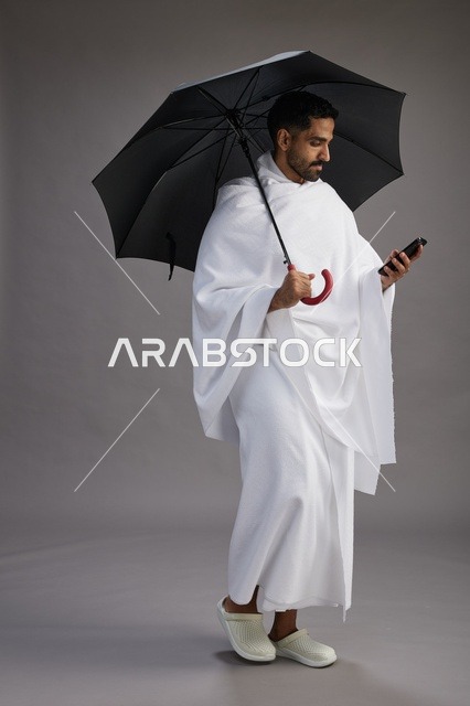 بورتريه لحاج  عربي سعودي  خليجي بملابس الإحرام يحمل مظله, الذهاب لاداء مناسك الحج والعمره, استخدام الهاتف المحمول  كامل الجسم, خلفيه رماديه