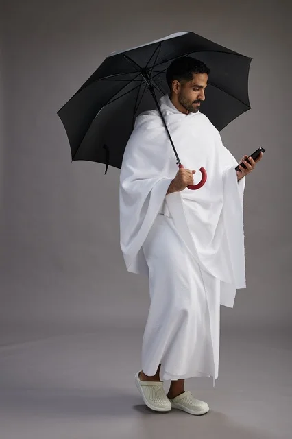 بورتريه لحاج  عربي سعودي  خليجي بملابس الإحرام يحمل مظله, الذهاب لاداء مناسك الحج والعمره, استخدام الهاتف المحمول  كامل الجسم, خلفيه رماديه