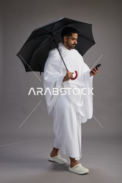 A portrait of an Arab Saudi Gulf pilgrim in Ihram clothing holding an umbrella, going to perform the rituals of Hajj and Umrah, using a mobile phone, full body, gray background.