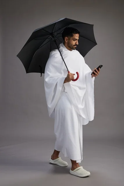 A portrait of an Arab Saudi Gulf pilgrim in Ihram clothing holding an umbrella, going to perform the rituals of Hajj and Umrah, using a mobile phone, full body, gray background. A portrait of an Arab Saudi Gulf pilgrim in Ihram clothing holding an umbrella, going to perform the rituals of Hajj and Umrah, using a mobile phone, full body, gray background.