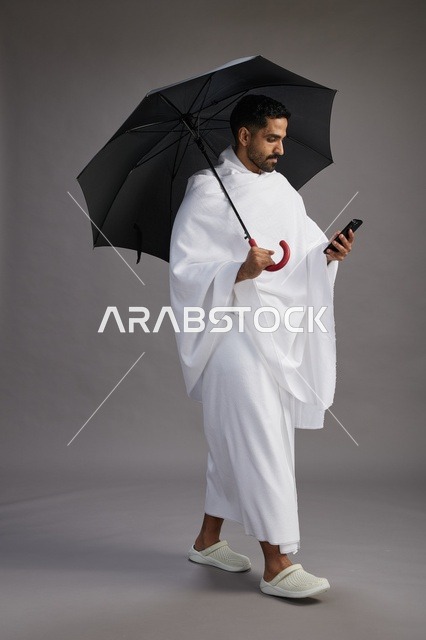 بورتريه لحاج  عربي سعودي  خليجي بملابس الإحرام يحمل مظله, الذهاب لاداء مناسك الحج والعمره, استخدام الهاتف المحمول  كامل الجسم, خلفيه رماديه