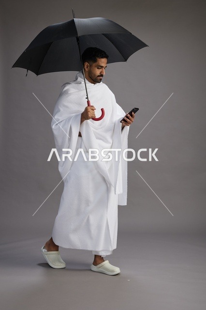 بورتريه لحاج  عربي سعودي  خليجي بملابس الإحرام يحمل مظله, الذهاب لاداء مناسك الحج والعمره, استخدام الهاتف المحمول  كامل الجسم, خلفيه رماديه