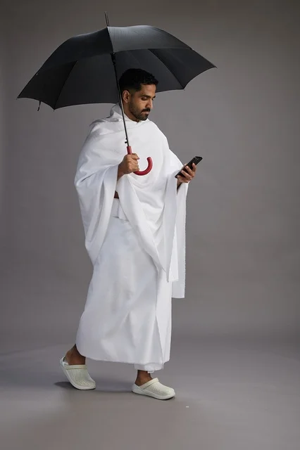A portrait of an Arab Saudi Gulf pilgrim in Ihram clothing holding an umbrella, going to perform the rituals of Hajj and Umrah, using a mobile phone, full body, gray background.