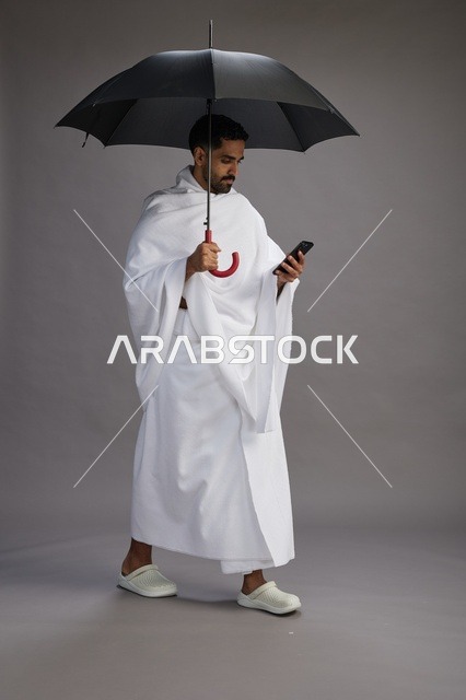 بورتريه لحاج  عربي سعودي  خليجي بملابس الإحرام يحمل مظله, الذهاب لاداء مناسك الحج والعمره, استخدام الهاتف المحمول  كامل الجسم, خلفيه رماديه