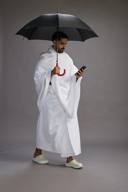 A portrait of an Arab Saudi Gulf pilgrim in Ihram clothing holding an umbrella, going to perform the rituals of Hajj and Umrah, using a mobile phone, full body, gray background.