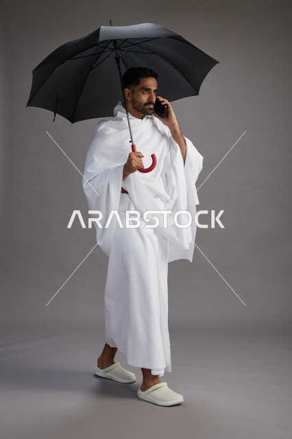بورتريه لحاج  عربي سعودي  خليجي بملابس الإحرام يحمل مظله, الذهاب لاداء مناسك الحج والعمره, استخدام الهاتف المحمول  كامل الجسم, خلفيه رماديه