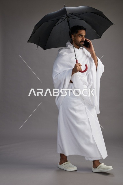 A portrait of an Arab Saudi Gulf pilgrim in Ihram clothing holding an umbrella, going to perform the rituals of Hajj and Umrah, using a mobile phone, full body, gray background.