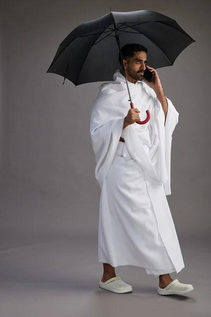 بورتريه لحاج  عربي سعودي  خليجي بملابس الإحرام يحمل مظله, الذهاب لاداء مناسك الحج والعمره, استخدام الهاتف المحمول  كامل الجسم, خلفيه رماديه