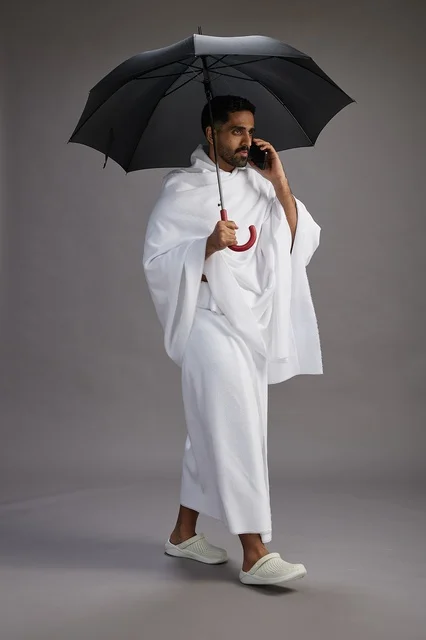 A portrait of an Arab Saudi Gulf pilgrim in Ihram clothing holding an umbrella, going to perform the rituals of Hajj and Umrah, using a mobile phone, full body, gray background.