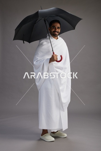 بورتريه لحاج  عربي سعودي  خليجي بملابس الإحرام يحمل مظله, الذهاب لاداء مناسك الحج والعمره,  كامل الجسم, خلفيه رماديه