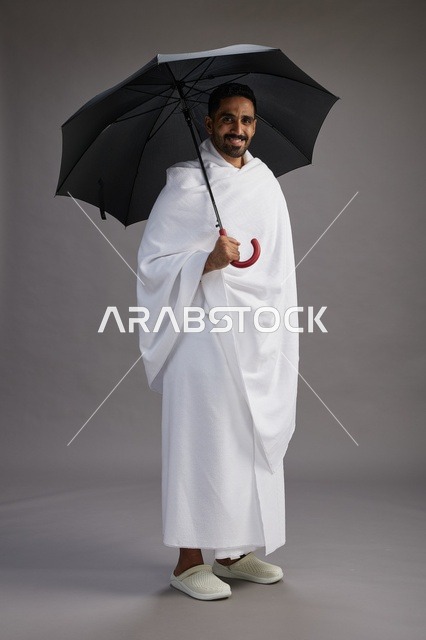 بورتريه لحاج  عربي سعودي  خليجي بملابس الإحرام يحمل مظله, الذهاب لاداء مناسك الحج والعمره,  كامل الجسم, خلفيه رماديه
