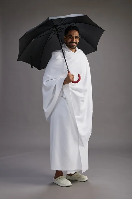 A portrait of an Arab Saudi Gulf pilgrim in Ihram clothing holding an umbrella, going to perform the rituals of Hajj and Umrah, full body, gray background.