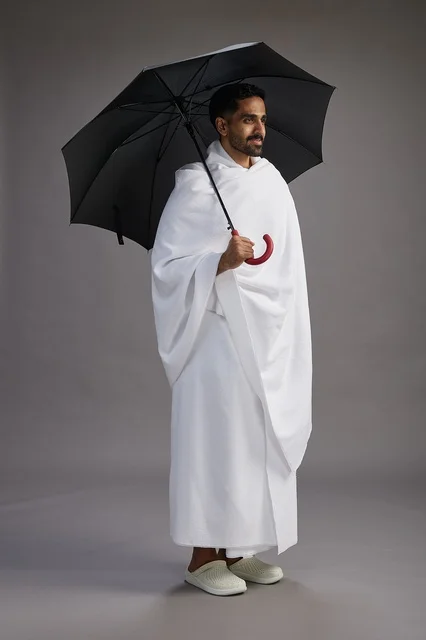 A portrait of an Arab Saudi Gulf pilgrim in Ihram clothing holding an umbrella, going to perform the rituals of Hajj and Umrah, full body, gray background.