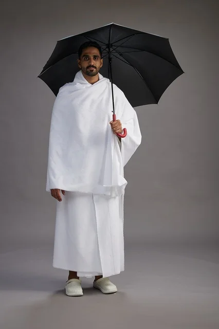 A portrait of an Arab Saudi Gulf pilgrim in Ihram clothing holding an umbrella, going to perform the rituals of Hajj and Umrah, full body, gray background.