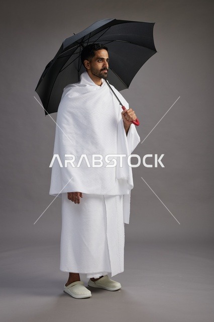 بورتريه لحاج  عربي سعودي  خليجي بملابس الإحرام يحمل مظله, الذهاب لاداء مناسك الحج والعمره,  كامل الجسم, خلفيه رماديه