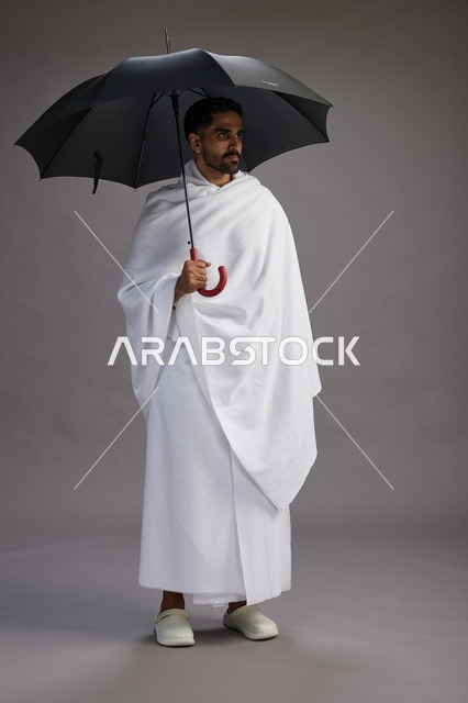 بورتريه لحاج  عربي سعودي  خليجي بملابس الإحرام يحمل مظله, الذهاب لاداء مناسك الحج والعمره,  كامل الجسم, خلفيه رماديه