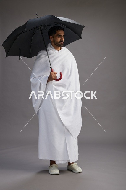 بورتريه لحاج  عربي سعودي  خليجي بملابس الإحرام يحمل مظله, الذهاب لاداء مناسك الحج والعمره,  كامل الجسم, خلفيه رماديه