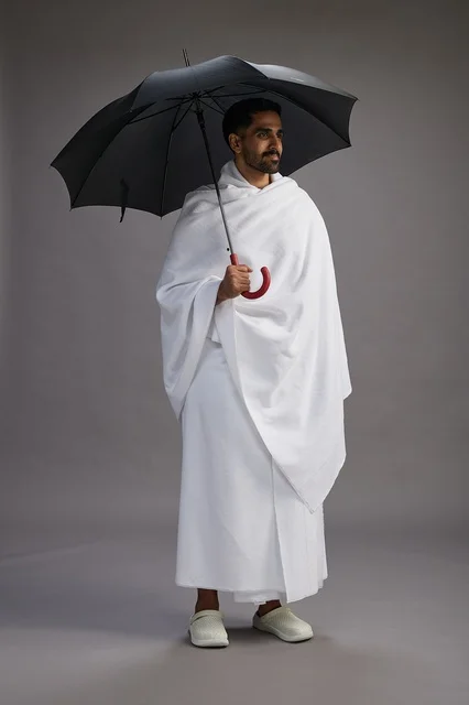 A portrait of an Arab Saudi Gulf pilgrim in Ihram clothing holding an umbrella, going to perform the rituals of Hajj and Umrah, full body, gray background.
