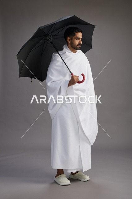 بورتريه لحاج  عربي سعودي  خليجي بملابس الإحرام يحمل مظله, الذهاب لاداء مناسك الحج والعمره,  كامل الجسم, خلفيه رماديه