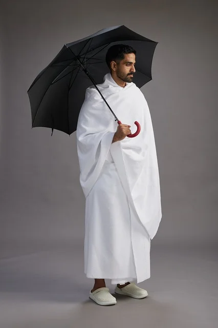 A portrait of an Arab Saudi Gulf pilgrim in Ihram clothing holding an umbrella, going to perform the rituals of Hajj and Umrah, full body, gray background. A portrait of an Arab Saudi Gulf pilgrim in Ihram clothing holding an umbrella, going to perform the rituals of Hajj and Umrah, full body, gray background.
