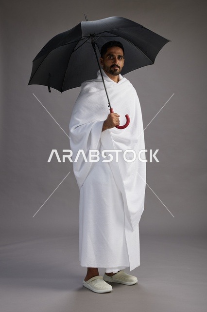 A portrait of an Arab Saudi Gulf pilgrim in Ihram clothing holding an umbrella, going to perform the rituals of Hajj and Umrah, full body, gray background.