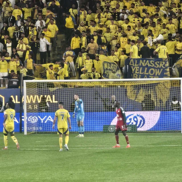 جماهير نادي النصر باللون الأصفر خلف المرمى في استاد الرياض، المملكة العربية السعودية، أجواء حماسية وهتافات مشجعة، مشهد رياضي يعكس شغف كرة القدم وروح التشجيع