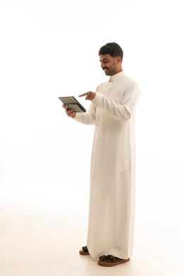 رجل سعودي يرتدي الثوب الأبيض يحمل جهاز لوحي ذكي واقفاً