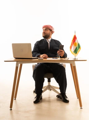 رجل كردي خلف المكتب مع حاسوب وعلم كردستان