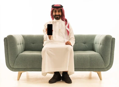 رجل سعودي يجلس على أريكة يحمل هاتفاً ذكياً بخلفية بيضاء
