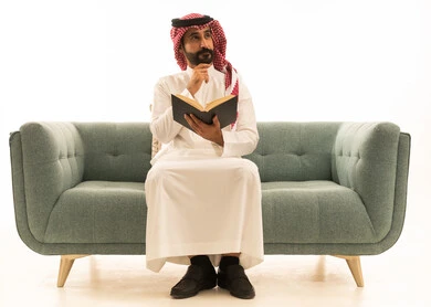 رجل سعودي يقرأ كتاباً على الأريكة بخلفية بيضاء