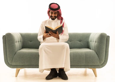 رجل سعودي باللباس التقليدي يقرأ كتاباً على أريكة خضراء