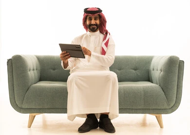 رجل سعودي مع جهاز لوحي على أريكة بخلفية بيضاء