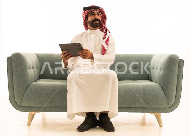 رجل سعودي مع جهاز لوحي جالس على أريكة بخلفية بيضاء