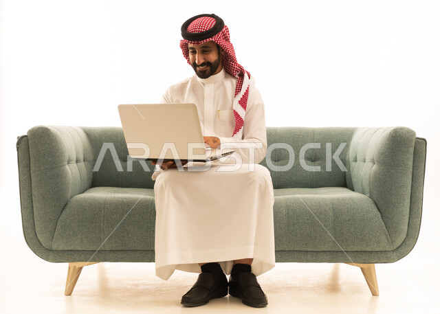رجل سعودي يستخدم اللابتوب على أريكة بخلفية بيضاء