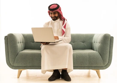 رجل سعودي يستخدم اللابتوب على أريكة بخلفية بيضاء