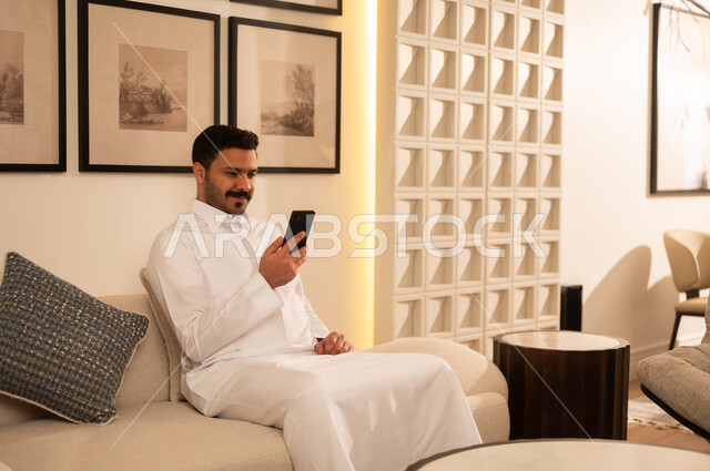 Saudi Man in Thobe Using Smartphone on Sofa
